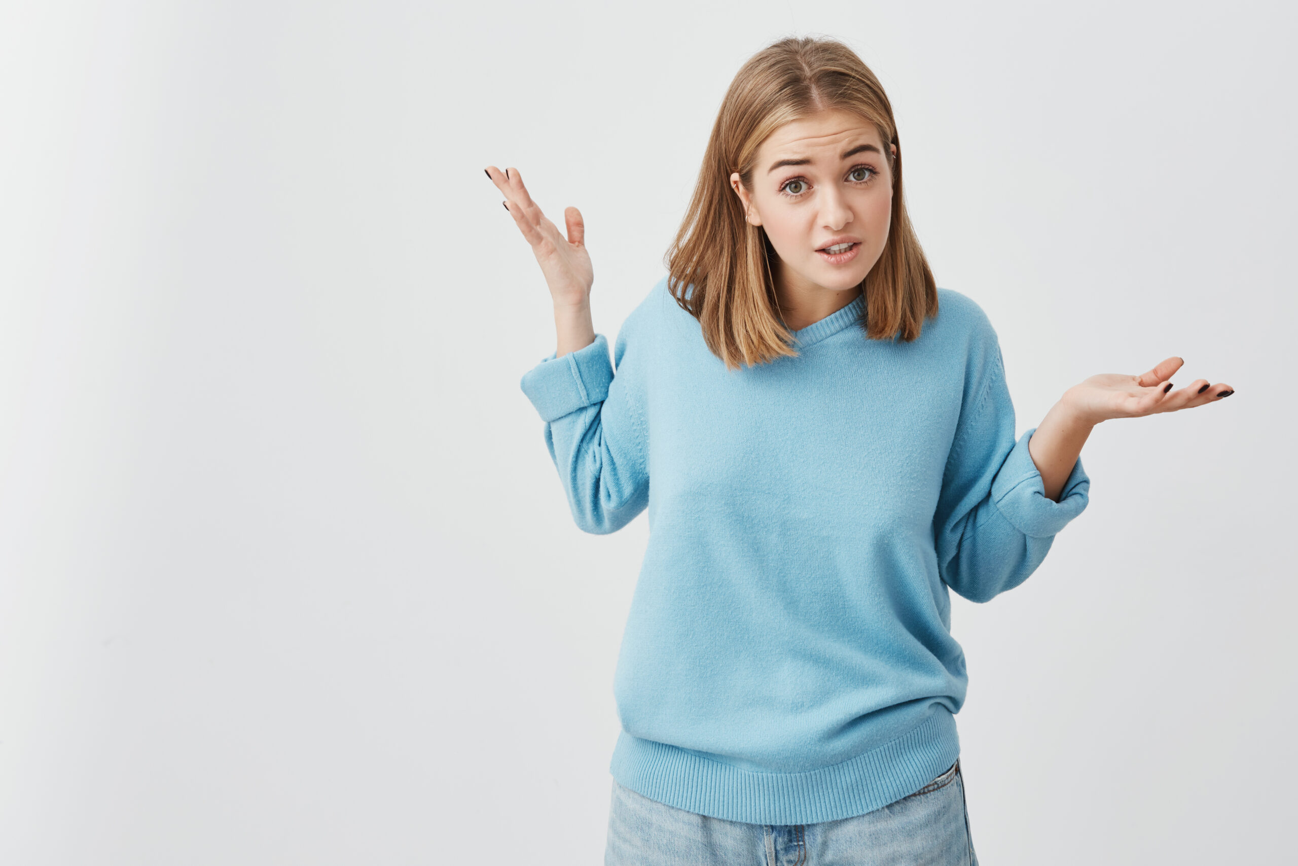 Clueless puzzled young pretty girl dressed in sweater and jeans shrugging shoulders and staring at camera with confused look after she did something wrong but not feeling sorry or guilty, not understanding what is wrong. Body language